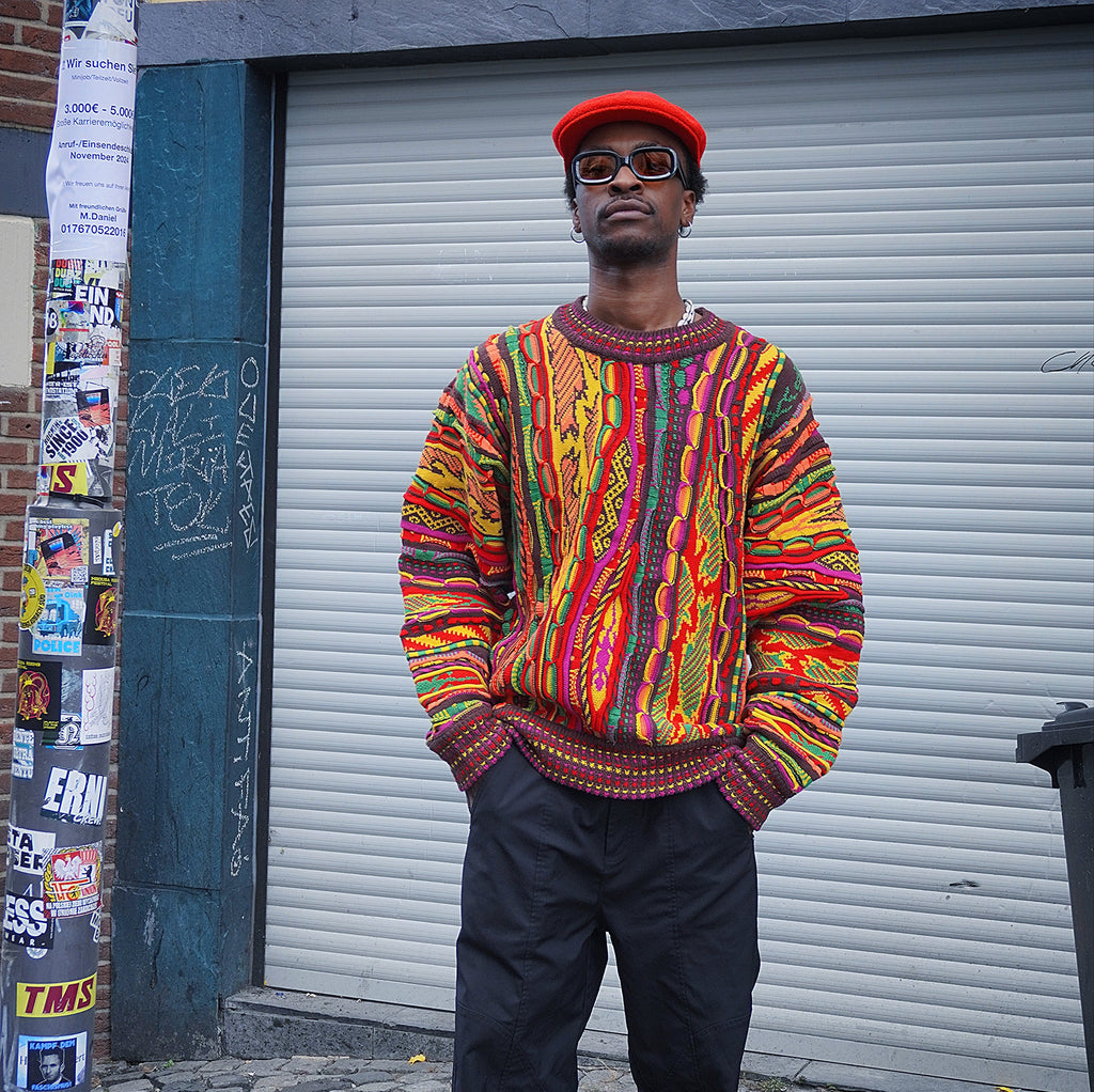 Model sitting on a graffiti wall wearing the multicolour 3D knit sweater with baggy light denim